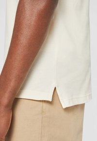 T-shirt à manches courtes de couleur crème avec un tissu texturé, fentes sur les côtés au niveau de l'ourlet, et coupe décontractée, associé à un pantalon beige.