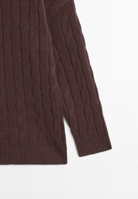 Manica di un maglione marrone scuro a trecce con polsino a coste, distesa su uno sfondo bianco.