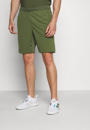Pantalones cortos deportivos verdes con una cintura elástica, con un detalle lateral en amarillo brillante. Combinados con zapatillas deportivas blancas.
