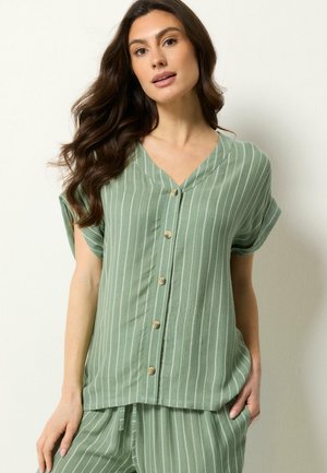 Vrouw met lang donker haar die een lichtgroene blouse met verticale strepen en bijpassende broek draagt, staand tegen een witte achtergrond.