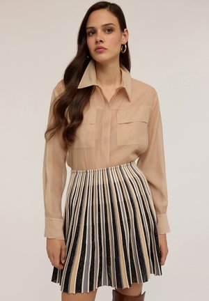 Blusa beige semitrasparente con tasche sul petto e colletto a punta abbinata a una gonna plissettata con righe verticali nere, beige e grigie.