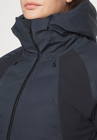 Veste bleu marine avec un col haut, présentant des sections de panneau noir contrastantes, une fermeture éclair sur le devant et des cordons de serrage ajustables au niveau de la capuche.