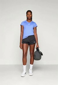 Světle modré sportovní tričko s krátkým rukávem, černé sportovní šortky a bílé tenisky. Drží malou černou gymnastickou tašku.