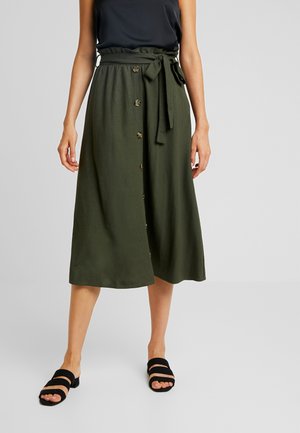 Mujer con falda midi verde oliva con botones delanteros y cinturón anudado en la cintura, combinada con top negro sin mangas y sandalias negras.