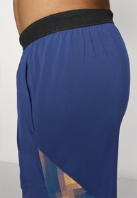 Blaue Sportshorts mit einem schwarzen elastischen Bund, ausgestattet mit einem seitlichen Mesh-Einsatz mit einem geometrischen Muster in Blau- und Gelbtönen.