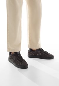 Sneakers in suede nera con accenti texturizzati, caratterizzati da un profilo basso, design con lacci e suole in gomma, abbinati a pantaloni crema.