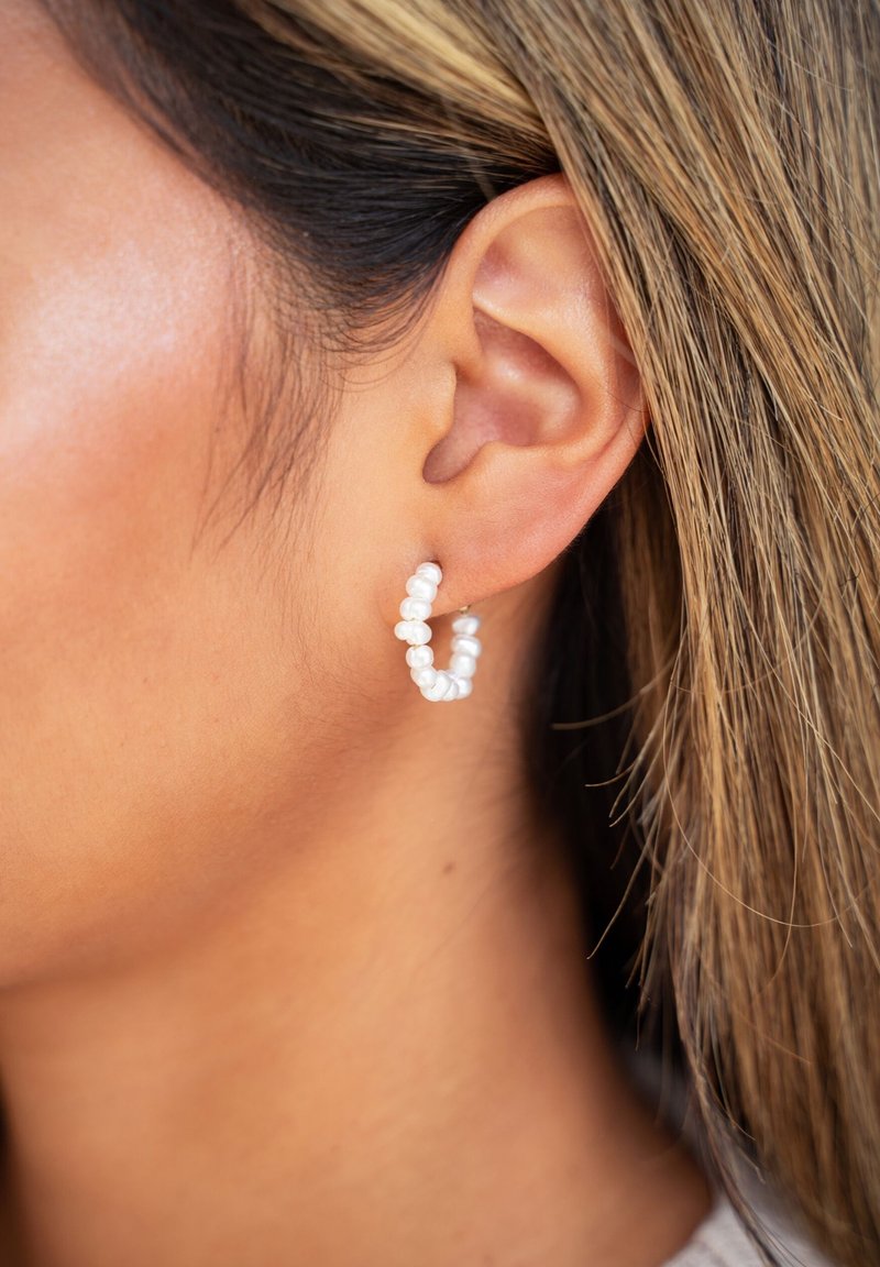Boucles d'oreilles en forme de créoles blanches en perles avec un design torsadé, présentant des formes irrégulières et une finition brillante. Une quincaillerie en ton argent sécurise les boucles d'oreilles.