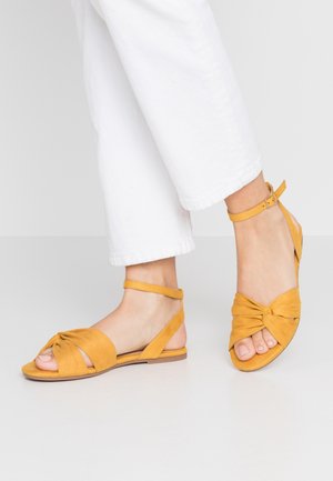 Yellow suede sandals with a knotted front design, open-toe style, and ankle strap. Set against white pants on a light background.
