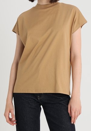 Woman wearing a plain tan short-sleeve t-shirt and dark jeans, standing against a light background.