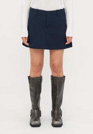 Navy blue skirt with front pleats and two side pockets, paired with knee-high gray leather boots featuring buckles and a textured finish.