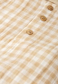 Tejido de mezcla en color beige y blanco con un patrón de cuadros y un acabado texturizado, adornado con cuatro botones de madera; costuras detalladas alrededor de la tapeta de botones.
