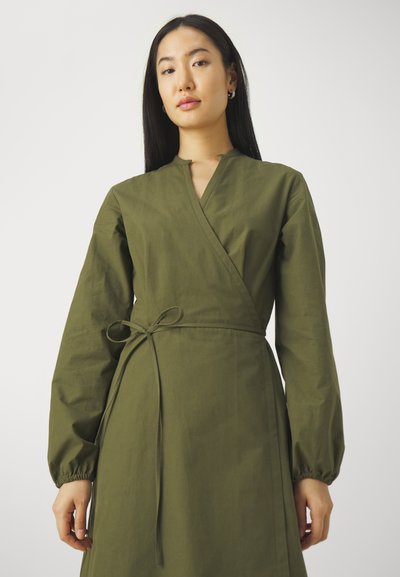 Robe portefeuille vert olive avec de longues manches bouffantes, des poignets élastiques et une ceinture à nouer à la taille. Fabriquée dans un tissu lisse et texturé.