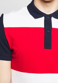 Polo avec des rayures horizontales rouge, blanche et bleu marine, col bleu marine et deux boutons. Tissu doux et texturé.