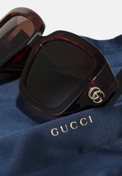 Gucci suurendusega prillid tumeda pruuni kilpkonnakuju raamiga, tumedate läätsede ja hõbedaste detailedega. Kaasas sinine riie, millel on kuldne "GUCCI".