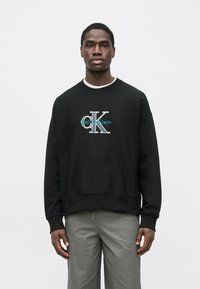Black sweatshirt with ribbed cuffs and hem, featuring a large white and turquoise "CK" logo. Cotton blend fabric, relaxed fit.