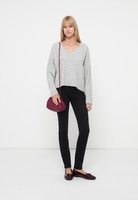 Suéter de pico gris claro, jeans skinny negros y bolso burdeos. La modelo lleva mocasines oscuros de charol y está de pie frente a un fondo blanco.