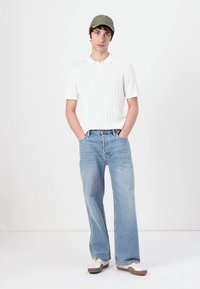 Hvid ribbet polo skjorte, lyseblå brede jeans og grøn kasket, parret med hvide sneakers med en brun sål. Afslappet outfit.