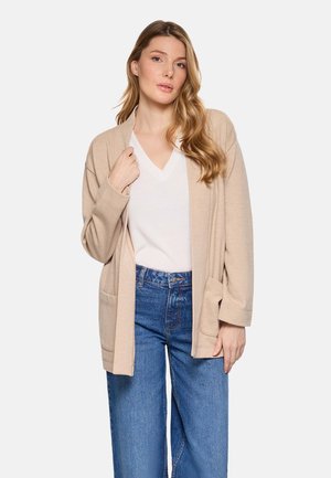Mujer con cabello largo y ondulado, vestida con un cárdigan beige, blusa blanca de cuello en V y jeans azules de talle alto, de pie frente a un fondo liso.
