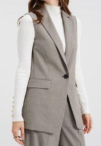 Kostkovaná vesta z hnědé a bílé tkaniny, na míru střižená, s jedním knoflíkem na zapínání, má boční kapsy a klopy s výřezem.