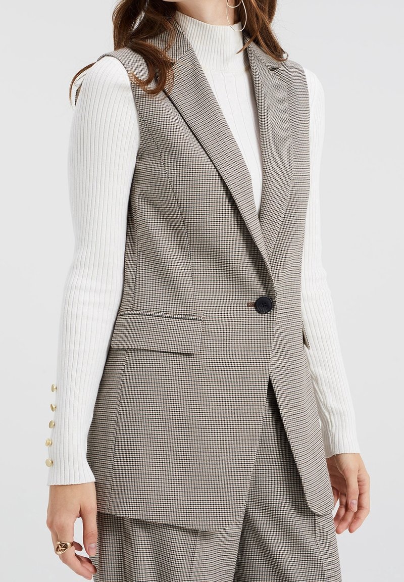 Kostkovaná vesta z hnědé a bílé tkaniny, na míru střižená, s jedním knoflíkem na zapínání, má boční kapsy a klopy s výřezem.