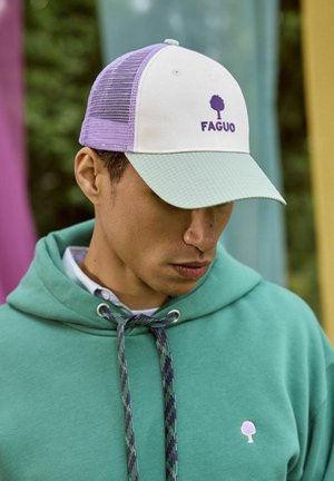 Jeune homme portant un sweat à capuche vert pastel et une casquette Faguo blanche et violette, regardant vers le bas en plein air.