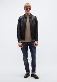 Fekete bőrkabát, bézs pulóver, kék farmer nadrág felhajtott szegéllyel és sötét cipő. A modell napszemüveget visel, egyszínű háttér előtt áll.