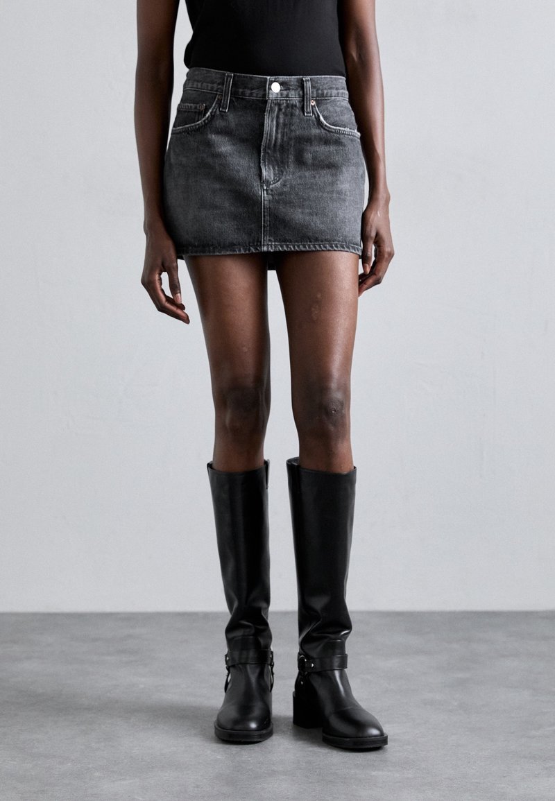 Personne portant une mini-jupe en jean gris foncé, un haut noir sans manches et des bottes noires à talons montant jusqu'aux genoux, debout sur un sol gris avec un fond uni.
