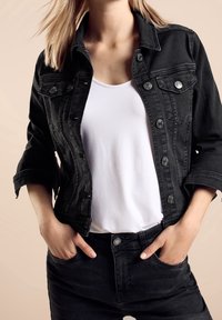 Svart denimjacka med silverknappar, figursydd design, uppvikta ärmar, lagd över en vit t-shirt, tillsammans med svarta jeans. Slät textur.