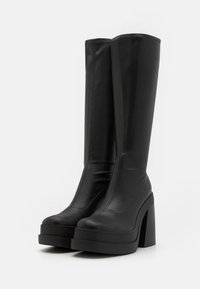 Bottes noires montant jusqu'aux genoux en cuir lisse avec une semelle plateforme épaisse et un talon bloc haut, avec des détails de couture visibles.