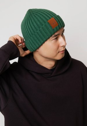 Zielona dzianinowa czapka typu beanie o prążkowanej fakturze z brązową skórzaną naszywką; noszona z ciemnofioletową bluzą z kapturem, częściowo zakrywająca ucho.