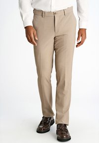 Pantaloni eleganti beige con una texture liscia, vestibilità su misura, passanti per cintura e chiusura con bottone frontale, abbinati a scarpe marroni con nappine.