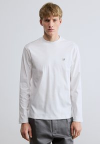 Fehér hosszú ujjú póló puha pamutból, kerek nyakkivágással és egy kis logóval a mellkason. Sima textúra és testhezálló szabás.