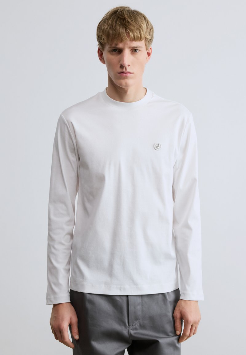 Fehér hosszú ujjú póló puha pamutból, kerek nyakkivágással és egy kis logóval a mellkason. Sima textúra és testhezálló szabás.