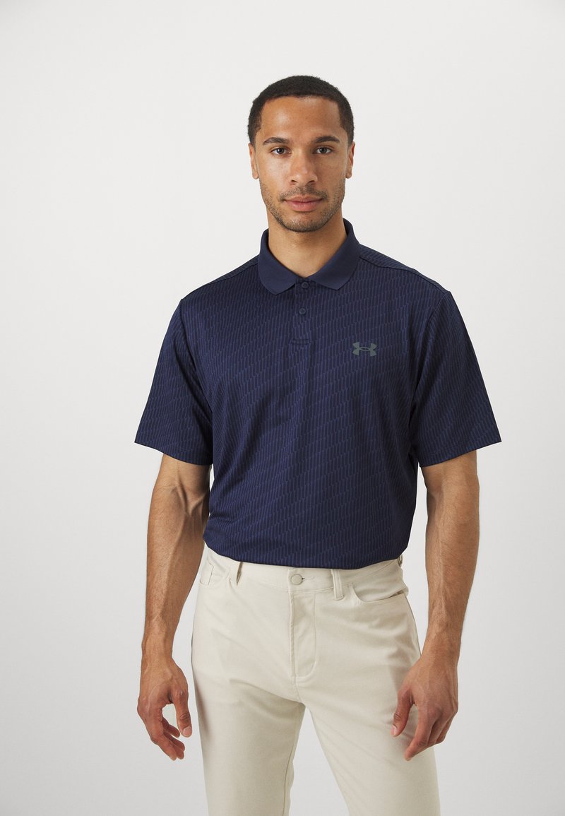 Polo de culoare bleumarin, cu un model geometric subtil, mâneci scurte și guler, asociat cu pantaloni bej deschis.