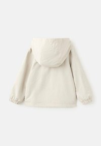 Svetlo bež jakna s kapuco, elastičnimi zapestniki in sproščenim krojem. Izdelana iz gladke tkanine z teksturirano površino. Brez vidnih zadrg.