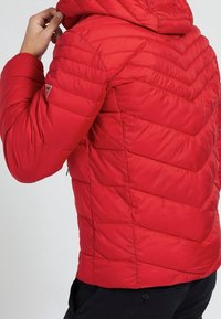 Rote isolierte Jacke mit einem Chevron-Steppdesign, verstellbarem Kapuze und einer seitlichen Reißverschlusstasche. Glatte Textur, leichtes Material.