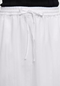 Pantalon blanc à fines rayures verticales avec une taille élastique nouée d'un nœud, associé à un haut noir.