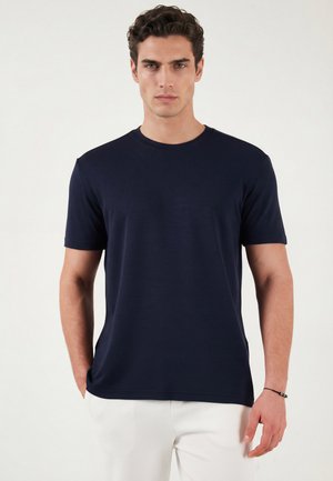 Mężczyzna o ciemnych, kręconych włosach, ubrany w granatowy t-shirt z krótkim rękawem i białe spodnie, stojący na jasnym tle.