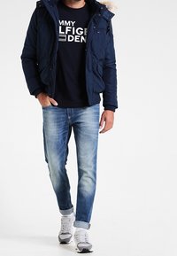 Man som bär marinblå parkas med pälsluva, svart Tommy Hilfiger-logot-sweatshirt, urtvättade blå jeans och vita sneakers går framåt.