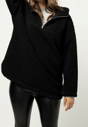 Kobieta ubrana w czarny polar z półzamekiem i czarne spodnie w skórzanym stylu, stojąca z uniesioną jedną ręką.