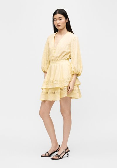 Γυναίκα που φοράει φόρεμα σε ανοιχτό κίτρινο με στρώσεις και φουσκωτά 3/4 μανίκια, με μαύρα σανδάλια με τακούνι και λουράκια, στέκεται μπροστά σε απλό φόντο.