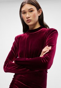 Μπορντό βελούδινο φόρεμα με μακριά μανίκια και ψεύτικο λαιμόκοψη, που διαθέτει μαζεμένες λεπτομέρειες στη μέση, λεία υφή και στενή σιλουέτα.