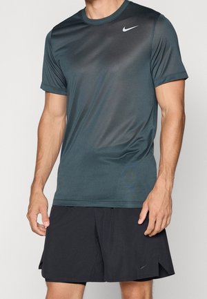 Mand iført mørkegrå Nike kortærmet sportstrøje og sorte Nike shorts står mod en ensfarvet baggrund.