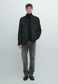 Giacca nera in misto lana con tasche oversize, indossata sopra un dolcevita nero e abbinata a jeans grigi in denim e mocassini neri.