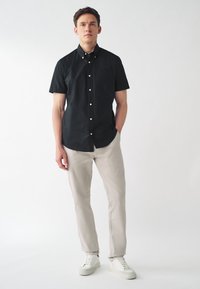 Camicia a maniche corte nera con bottoni bianchi, abbinata a pantaloni beige chiaro e sneaker bianche. Design semplice, senza fantasie.