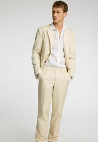 Ljusbeige kostym med en skräddarsydd kavaj med svarta knappar, andningsbar vit skjorta och byxor med lös passform och elastisk midja.