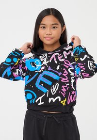 Giovane ragazza che aggiusta una felpa con cappuccio in stile graffiti colorato, con motivi neri, blu, rosa, bianchi e gialli, indossando pantaloni neri con cordino, su uno sfondo neutro.