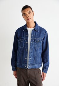 Jonge persoon in een blauwe spijkerjasje over een wit shirt en een broek in een bruine kleur, staande voor een effen witte achtergrond.