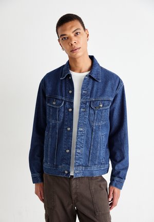 Young person wearing a blue denim jacket over a white shirt and brown pants, standing against a plain white background.