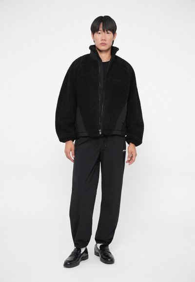 Zwarte faux fur zip-up jas met een hoge kraag, gecombineerd met losse zwarte broeken en zwarte instapschoenen. Eenvoudig, monochroom ontwerp.